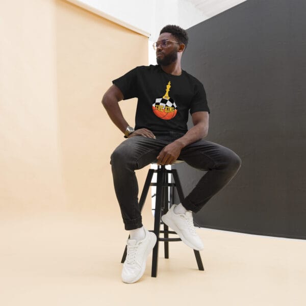 Man sitting down wearing classic tee in black with text 'The King', a basketball with top half a chess board and King piece on top with number 23