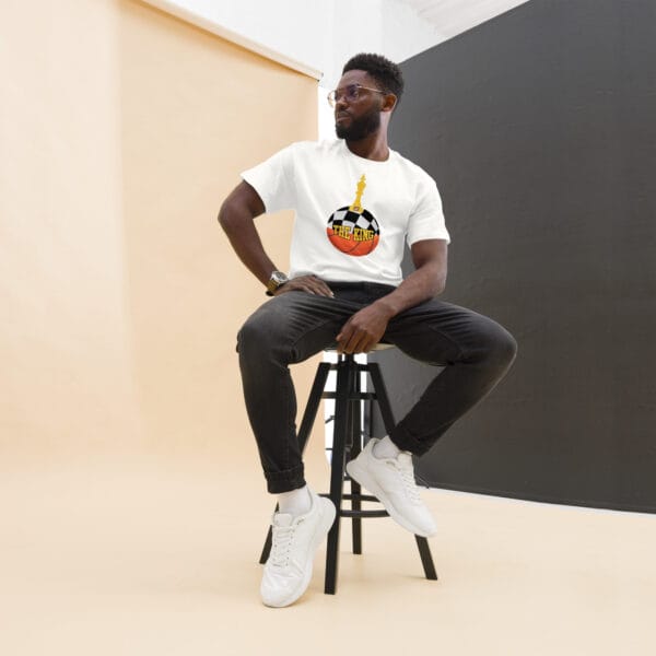 Man sitting down wearing classic tee in white with text 'The King', a basketball with top half a chess board and King piece on top with number 23