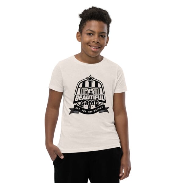 Boy in a heather dust staple t-shirt with a black The Beautiful Game crest, est. for the people